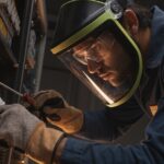 Male electrician wearing protective face shield and safety goggles near high-voltage panel.