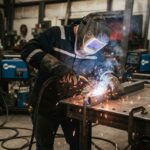 A male welder wearing a flame resistant jacket and helmet grinding metal in a workshop.
